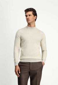 Jasno beżowy sweter z okrągłym dekoltem, ściągaczami przy mankietach i na dolnym wykończeniu. Materiał wydaje się gładki i lekko teksturowany, noszony na brązowych dopasowanych spodniach.