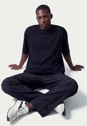 Uomo seduto a gambe incrociate sul pavimento, indossa una maglietta nera, pantaloni a righe sottili e sneakers bianche, con le mani appoggiate dietro di sé.