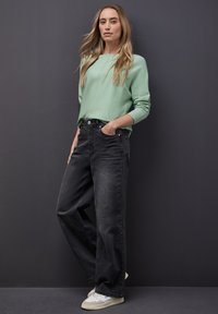 Hellgrüner Strickpullover mit langen Ärmeln, kombiniert mit hoch sitzenden, schwarzen Denim-Jeans. Weiße Turnschuhe ergänzen das Outfit.