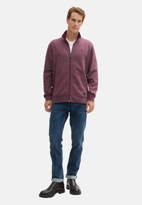 Felpa con zip color bordeaux e colletto alto, con due tasche frontali, abbinata a jeans blu e stivali neri. Texture liscia, design casual.