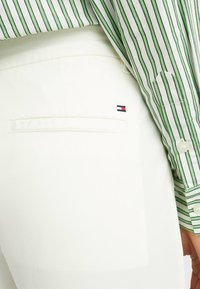 Pantaloni bianchi con dettaglio sulla tasca posteriore che presenta un piccolo emblema blu, rosso e bianco. Camicia a righe verde e bianca parzialmente visibile.
