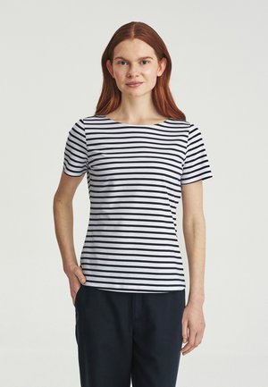 Jeune femme aux longs cheveux roux portant un t-shirt à manches courtes blanc et noir à rayures horizontales et un pantalon foncé, debout, une main dans la poche.
