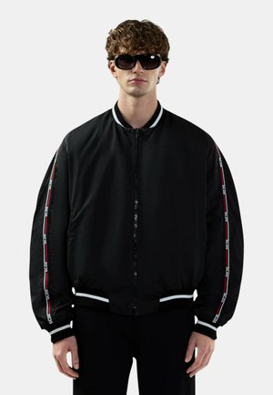 Giovane uomo che indossa una giacca bomber nera con polsini a righe bianche e marchio "GCDS" sulle maniche, abbinata a grandi occhiali da sole neri.