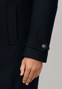 Mano accanto a una manica di un cappotto di lana blu notte con dettagli di fibbia e tasca a pattina su uno sfondo neutro.