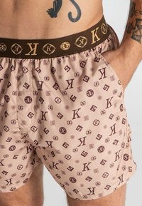 Pantaloni corti rosa chiaro con un motivo a logo ripetuto in marrone scuro. Presentano una vita elastica marrone con dettagli del logo e cuciture laterali.
