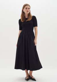 Schwarzes Kleid mit tailliertem Oberteil und kurzen Ärmeln, das eine geraffte Taille und einen voluminösen Rock aufweist. Glattes Gewebe mit matter Oberfläche.