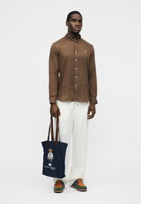 Bărbat purtând o cămașă maro cu nasturi și pantaloni albi, ținând o geantă de umăr albastru închis cu un desen cu urs, stând în fața unui fundal alb simplu.