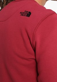 Sudadera de forro polar rojo con cuello redondo, tejido texturizado y un logo negro en el hombro izquierdo. Acabado suave con costuras visibles.