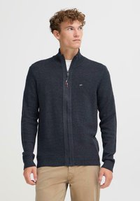 Tamsiai pilka zip-up megztinis su tekstūruotu mezgimo raštu, aukštu apykakle ir raibomis rankovėmis; turi mažą logotipą ant krūtinės.