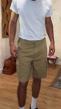 Homme portant un t-shirt blanc et un short cargo kaki, debout à l'intérieur sur un sol en bois près d'un sac en cuir marron et d'une petite table avec des livres.