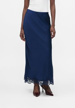 Frau trägt einen marineblauen Satin-Midirock mit gewelltem Spitzen Saum und bordeauxroten spitz zulaufenden High Heels, stehend vor weißem Hintergrund.
