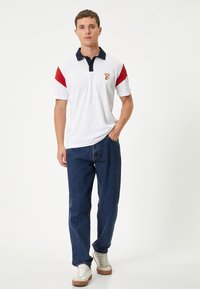 Polo skjorte i hvid med røde og marineblå detaljer, med korte ærmer, en krave og et lille broderet logo. Båret med blå jeans og sneakers.