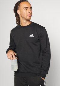 Felpa nera con scollo rotondo, realizzata in un materiale morbido. Presenta un logo bianco Adidas sul lato sinistro del petto. Design slim fit.