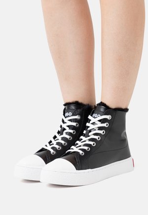 Baskets montantes noires avec lacets et semelles blanches, doublure en fourrure en haut, portées sur des jambes nues sur un fond uni.