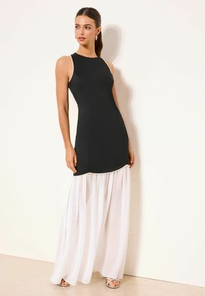 Nő ujjatlan fekete testhezálló ruhában, gyöngydísszel és lenge fehér szoknyával, egy egyszerű világos fal előtt állva.