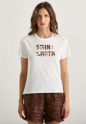 Donna con capelli castani mossi che indossa una t-shirt bianca con la scritta "SAINT BARTH" in stampa leopardata e pantaloncini marroni con stampa leopardata, in piedi contro uno sfondo semplice.