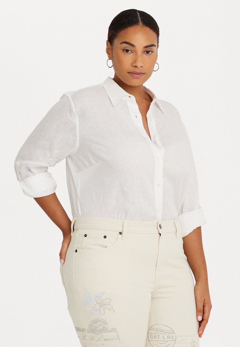 Camisa blanca de lino con botones y mangas enrolladas, combinada con pantalones beige claros que presentan acentos de estampados florales en azul y motivos postales.