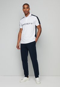 Polo bianco con accenti blu navy e logo "HACKETT". Abbinato a pantaloni della tuta blu navy e sneakers bianche. Tessuto morbido, maniche corte.