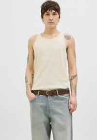 Canotta a coste color crema con scollatura rotonda, abbinata a jeans azzurri chiari e una cintura marrone. Mostra tatuaggi sulle braccia; vestibilità casual.