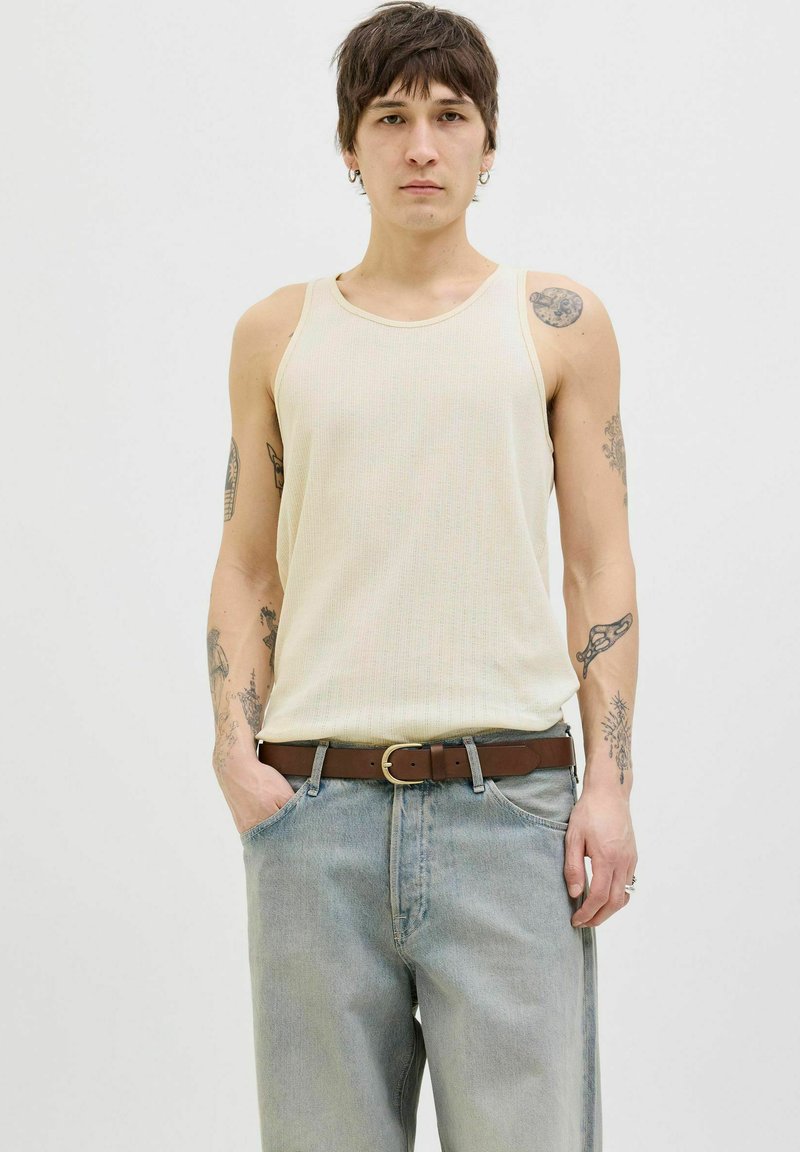 Canotta a coste color crema con scollatura rotonda, abbinata a jeans azzurri chiari e una cintura marrone. Mostra tatuaggi sulle braccia; vestibilità casual.