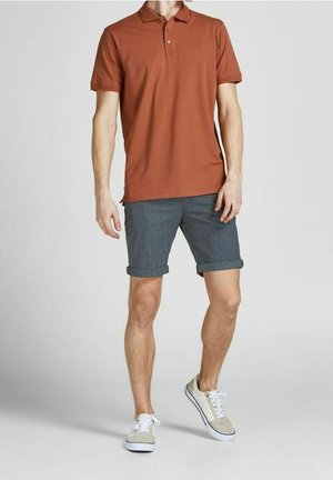 Homme portant un polo couleur rouille, un short gris foncé retroussé et des baskets beiges, debout devant un fond uni.