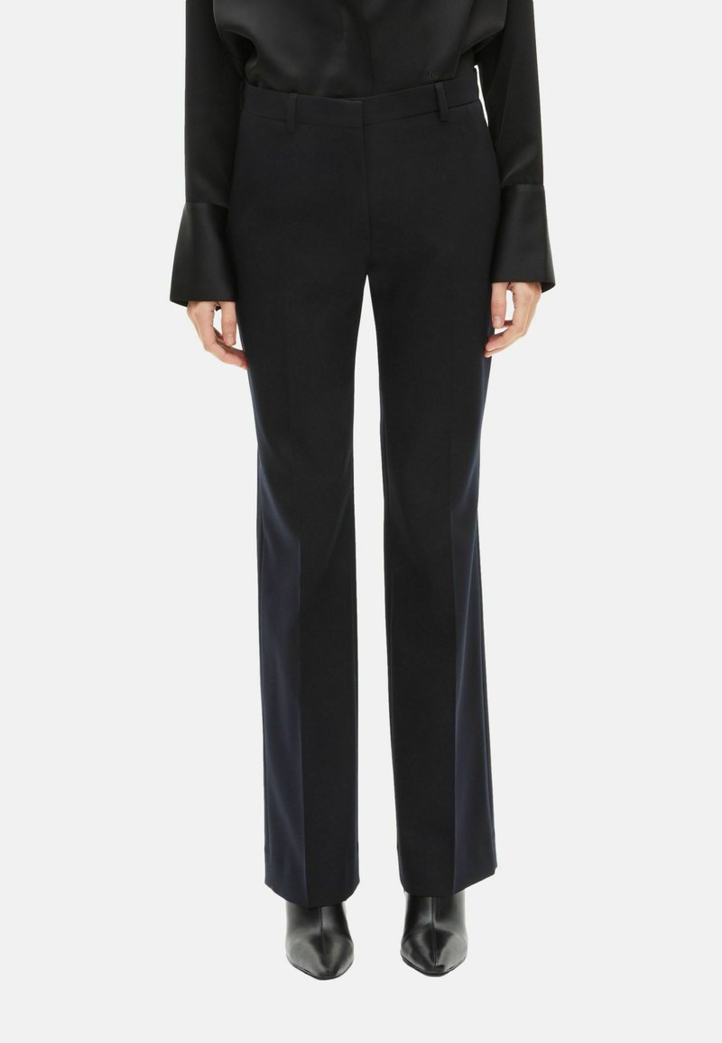 Schwarze, maßgeschneiderte Hosen mit geradem Bein, die eine glatte Textur und dezente Seitenakzente aufweisen. Kombiniert mit eleganten schwarzen Absatzschuhen.