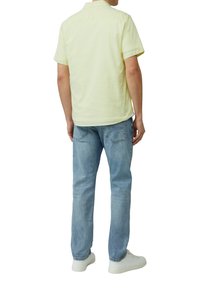 Helles gelbes kurzärmliges Hemd mit Kragen, kombiniert mit hellblauen Jeans und weißen Sneakers, von hinten betrachtet.
