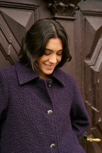 Cappotto di lana viola con texture e colletto, dotato di grandi bottoni neri e una vestibilità comoda, in contrasto con uno sfondo di una porta di legno scuro.