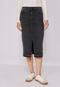 Hame midi denim hitam dengan belahan depan, tepi kasar, dan desain lima kantong. Dipasangkan dengan sepatu sneaker putih dan atasan lengan panjang berwarna terang.