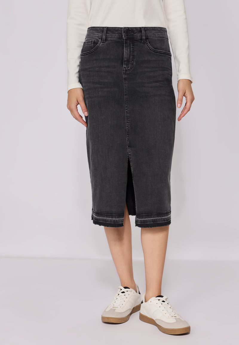 Hame midi denim hitam dengan belahan depan, tepi kasar, dan desain lima kantong. Dipasangkan dengan sepatu sneaker putih dan atasan lengan panjang berwarna terang.
