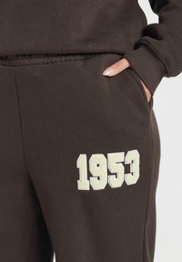 Bruna sweatpants med elastisk midja, sidficka och en krämfärgad texturerad tryck av året "1953" på låret.