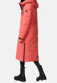 Abrigo largo de coral tipo puff con capucha, con un diseño acolchado, cremalleras laterales y parche negro con logo. Lleva puesto con botas de tobillo negras y pantalones.