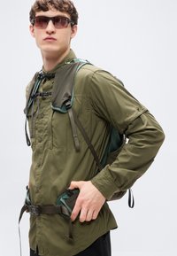 Καφέ χακί πουκάμισο με μακριά μανίκια, κλείσιμο με κουμπιά, τσέπες στο στήθος και ρυθμιζόμενα λουράκια. Φορεμένο με μαύρα γυαλιά ηλίου και σακίδιο πλάτης με πράσινες λεπτομέρειες.