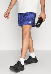 Ανδρικά αθλητικά σορτς σε μοβ με κυματιστό μοτίβο, σε συνδυασμό με μαύρα παπούτσια και λευκές κάλτσες αστραγάλου. Κρατάει ένα μαύρο μπουκάλι νερού.