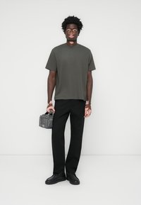 Olivengrünes T-Shirt mit kurzen Ärmeln, schwarze gerade Hosen, schwarze Schuhe, hält eine graue Tasche mit Reißverschluss. Einfaches Design, keine Muster.