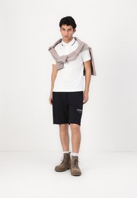 Hvit polo-skjorte med svarte detaljer, svarte shorts og beige ankelstøvler. En grå genser ligger over skuldrene. Uformell design.