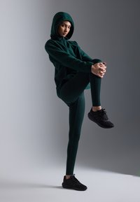 Grønn hettegenser med hette, kombinert med matchende grønne tights. Svarte joggesko. Modellen poserer med ett ben hevet, minimalistisk bakgrunn.
