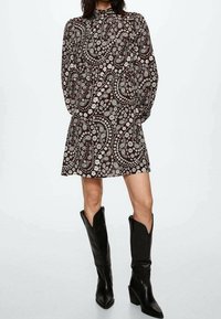 Braun gemustertes Kleid mit Paisleymuster, langen Ärmeln, hohem Ausschnitt und schwingendem Rock, kombiniert mit kniebedeckenden schwarzen spitzen Stiefeln.