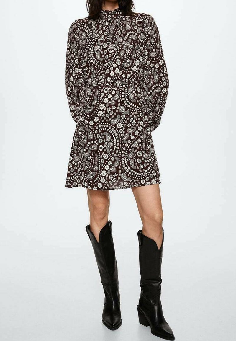 Braun gemustertes Kleid mit Paisleymuster, langen Ärmeln, hohem Ausschnitt und schwingendem Rock, kombiniert mit kniebedeckenden schwarzen spitzen Stiefeln.