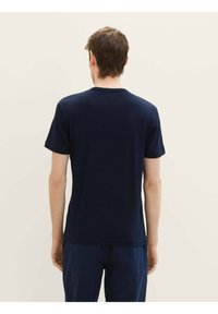 T-shirt bleu foncé à manches courtes, col rond et texture lisse. Design simple sans logos ni motifs visibles. Porté avec un pantalon.