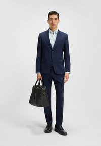 Completo su misura blu scuro con un motivo a quadri, camicia bianca, scarpe nere, in mano una borsa in pelle nera strutturata. Design elegante e definito.
