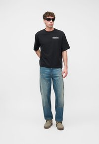 Νεαρός άνδρας με μαύρο T-shirt Timberland, μπλε τζιν με τσέπες θέσης, μπεζ μποτάκια και μαύρα γυαλιά ηλίου, στέκεται μπροστά σε απλό φόντο.