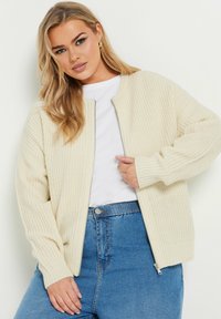 Beige stickad cardigan med dragkedja fram, ribbad manschett och avslappnad passform, bärs över en vit skjorta och blå högmidjade jeans.
