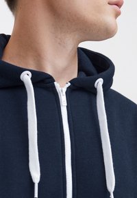 Sudadera con cremallera en color azul marino, con una cremallera y cordones blancos. Tela suave y un corte relajado, con un bolsillo tipo canguro en la parte inferior.