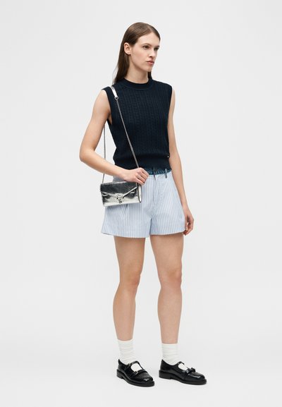 Νέα γυναίκα που φοράει μαύρο τοπ χωρίς μανίκια, ριγέ σορτς, λευκές κάλτσες, μαύρα μοκασίνια και κρατάει μια μικρή ασημένια τσάντα χιαστί.