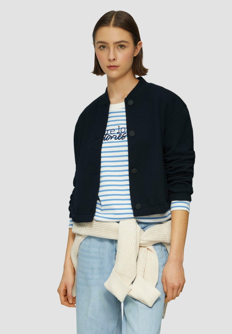 Veste bomber navy avec col rond, fermeture à boutons et manches longues ; superposée sur une chemise rayée bleu et blanc ; jeans bleu clair avec un pull crème noué à la taille.