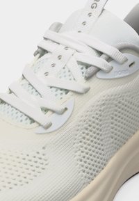 Κοντινό πλάνο λευκού πλεκτού αθλητικού παπουτσιού με γλώσσα από δίχτυ, λευκά επίπεδα κορδόνια που περνούν μέσα από υφασμάτινους κρίκους και ορατό άκρο ενισχυμένης σόλας.