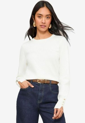 Weißer Langarmpullover mit Rundhalsausschnitt und geknöpften Manschetten, kombiniert mit dunkelblauen hoch-taillierten Jeans und einem braunen Gürtel.