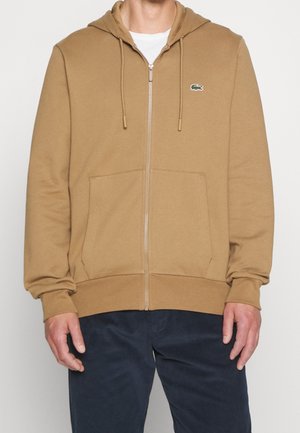 Hombre con sudadera con capucha marrón claro con cremallera, bolsillos delanteros y un pequeño logo verde de cocodrilo en el pecho izquierdo, combinado con pantalones oscuros.