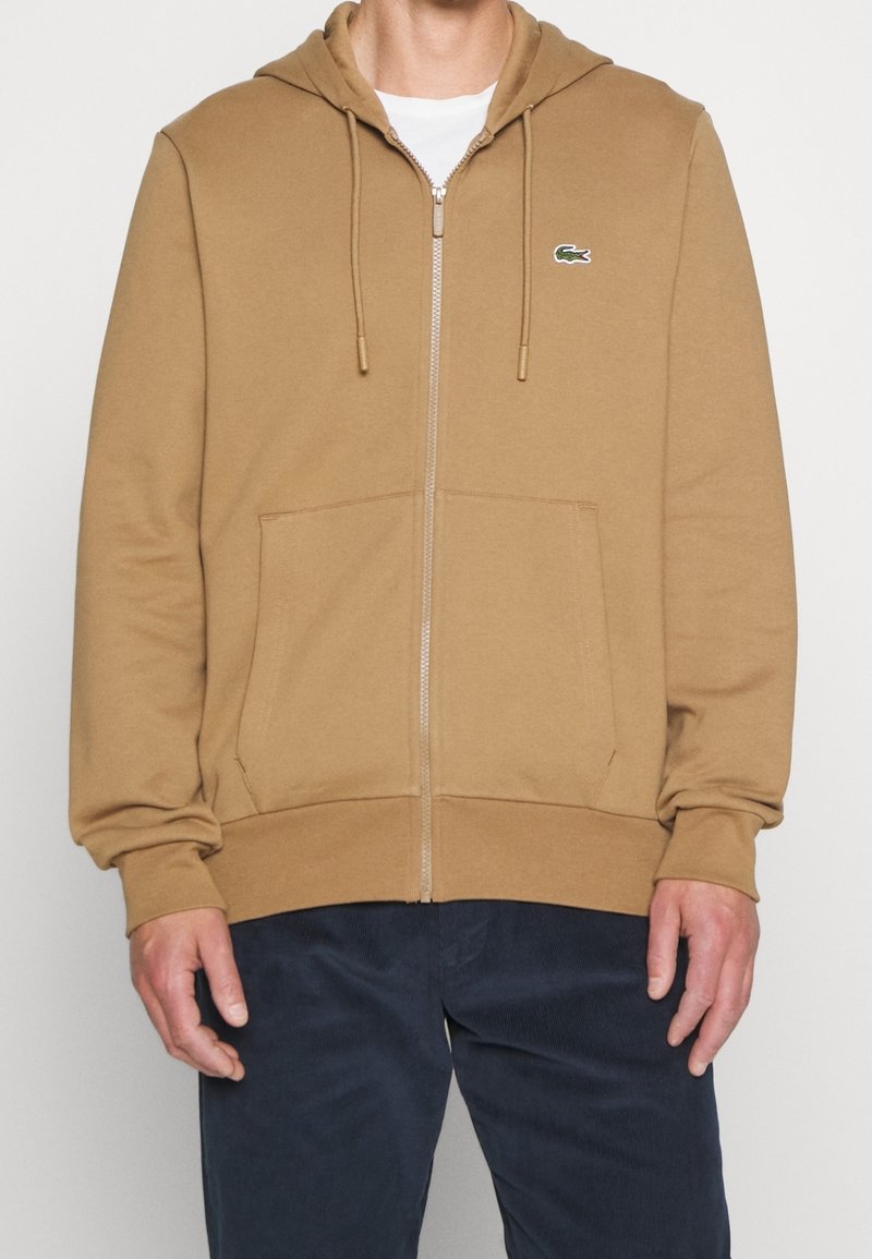 Homme portant un sweat à capuche beige zippé avec des poches avant et un petit logo crocodile vert sur la poitrine gauche, associé à un pantalon foncé.